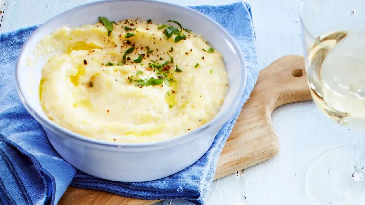 Cremige Polenta, verfeinert mit Butter und Kräutern, serviert in einer Schüssel. Ein Glas Weißwein und ein Holzbrett vervollständigen dieses Polenta Rezept.