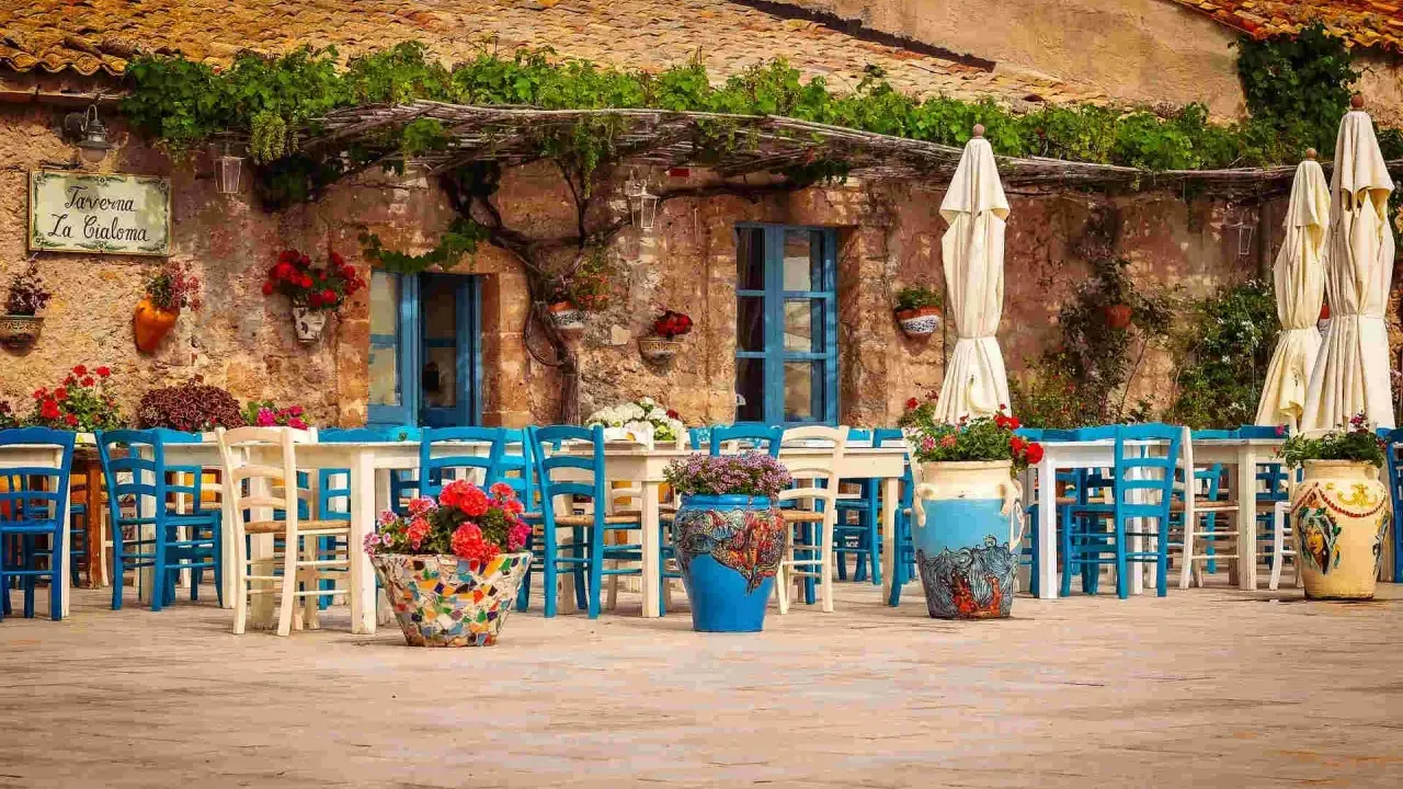 Taverna La Cialoma, un angolo incantevole di Siracusa e dintorni, con tavoli apparecchiati e sedie blu sotto un pergolato fiorito.