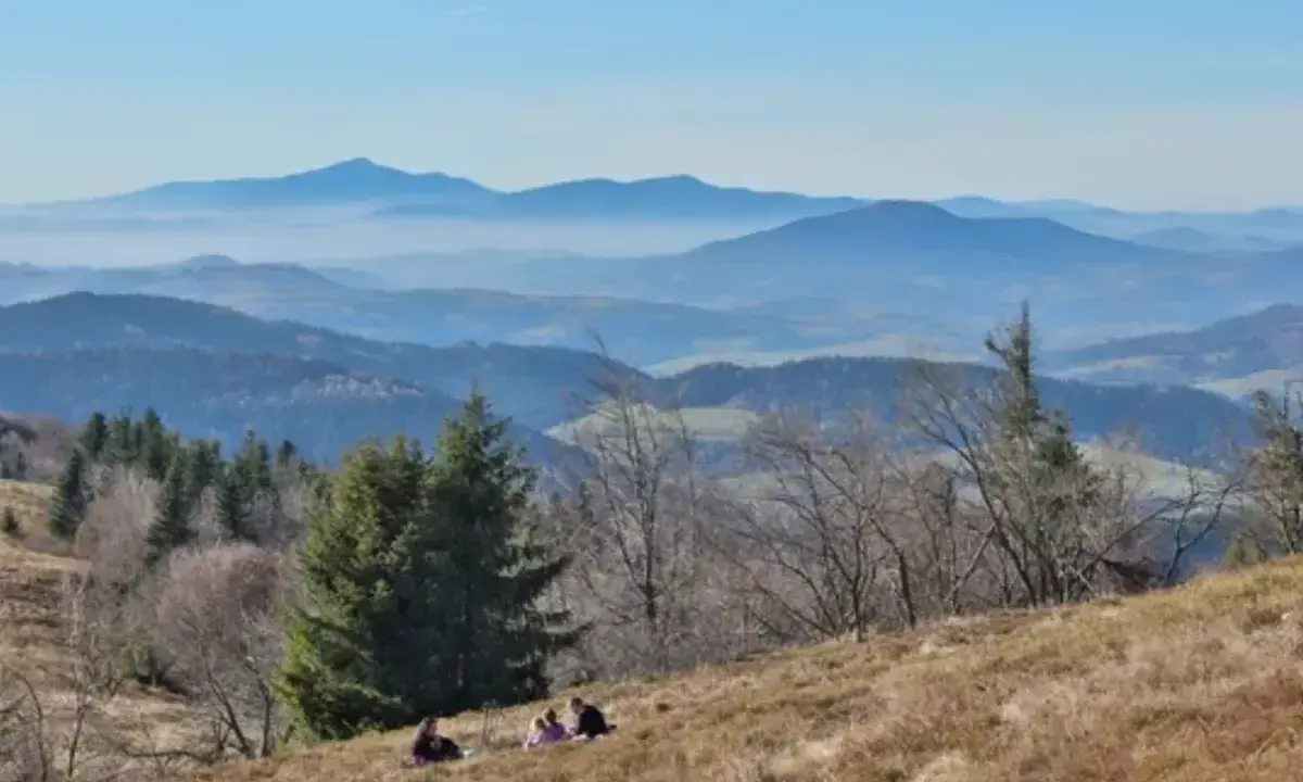 Widok na Beskid Wyspowy z kilkoma osobami odpoczywającymi na zboczu. W oddali widać kolejne pasma górskie w lekkiej mgle.