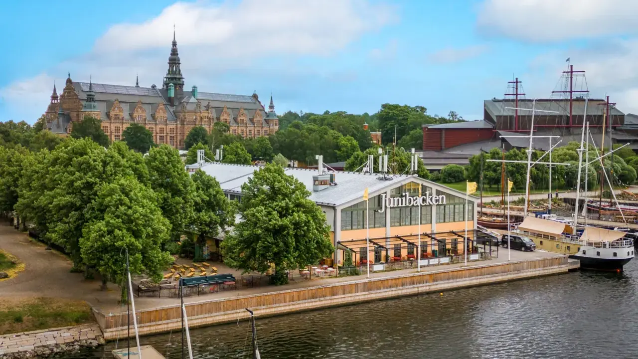 Junibacken, ett museum tillägnat Astrid Lindgren, ligger vid vattnet i Stockholm, med en stor byggnad i bakgrunden och segelfartyg i hamnen.