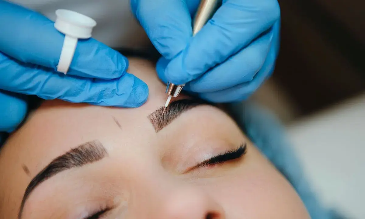 Main d'une esthéticienne en bleu, effectuant un entretien microblading sur les sourcils d'une cliente.