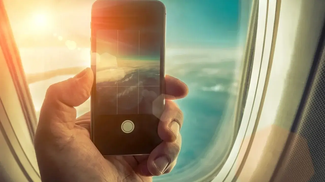 Dłoń trzyma smartfon, fotografując chmury z okna samolotu. Czy w samolocie można używać telefonu? Tak, do robienia zdjęć!