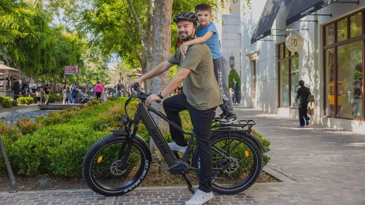 Ojciec z synem na rowerze elektrycznym, który wygląda jak jazda rowerem elektrycznym bez wspomagania. Słońce, zieleń i sklepy w tle.