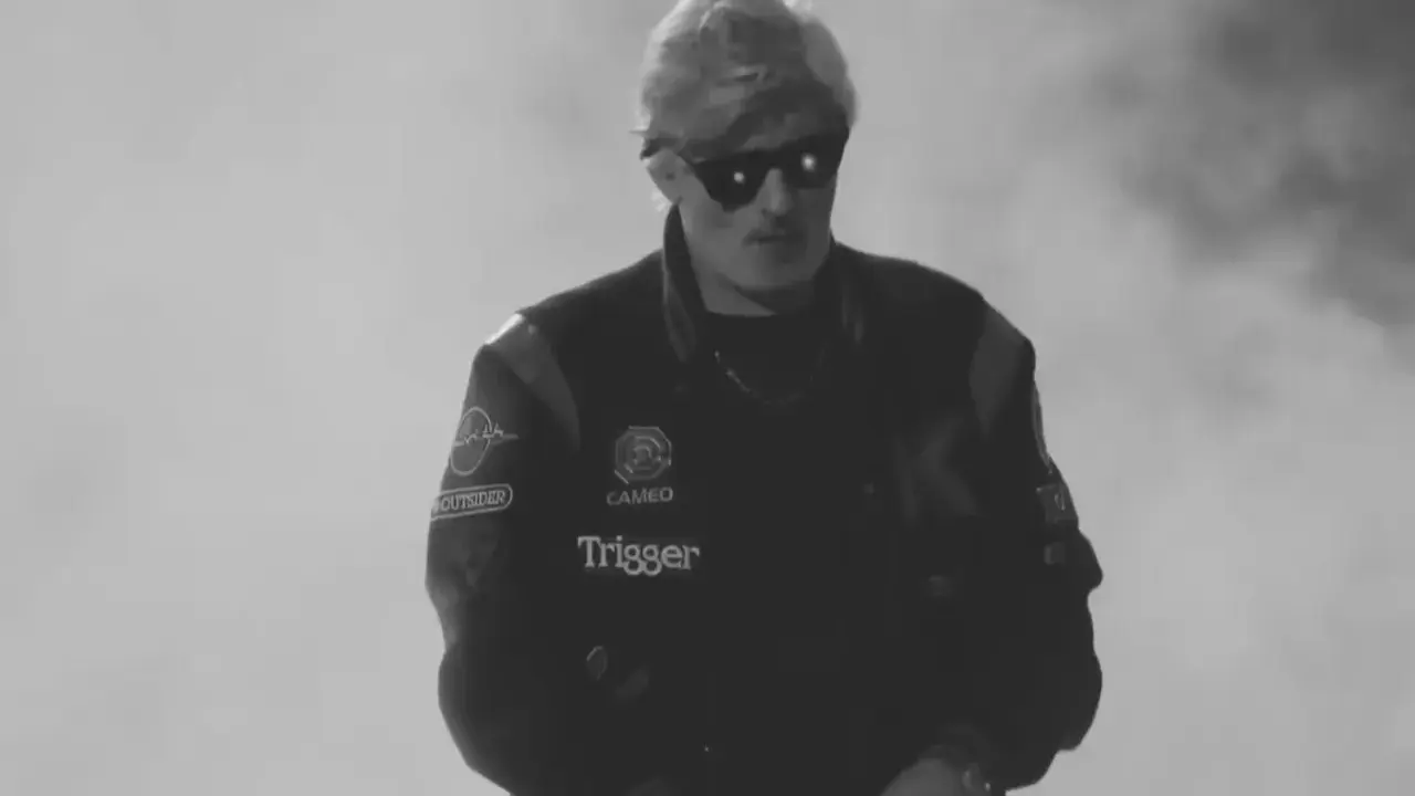 Młody artysta w okularach przeciwsłonecznych, w kurtce z naszywkami, gotowy do występu. Jego styl to czysty ras hip-hop.