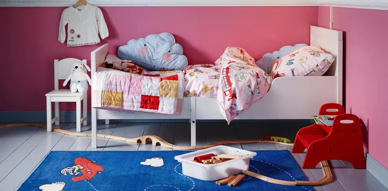 Habitación infantil con dos camas blancas, cojines de nube y mantas coloridas. Alfombra azul con tren de juguete y vías.