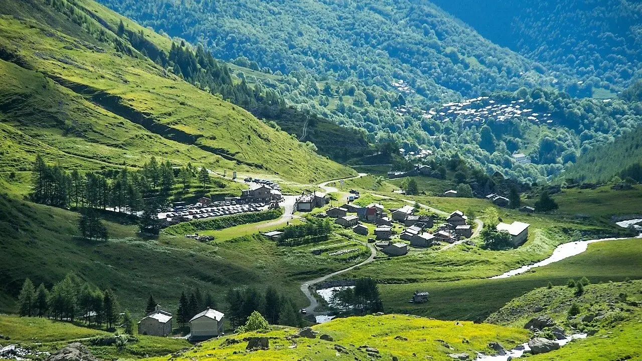 Un villaggio alpino nel **Pian della Regina**, con case in pietra e un fiume che scorre tra prati verdi e boschi lussureggianti.