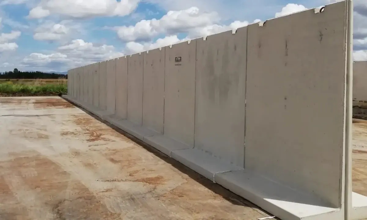 Larga pared de separador de hormigón prefabricado, instalada sobre una base de hormigón, con un cielo azul y nubes blancas al fondo.