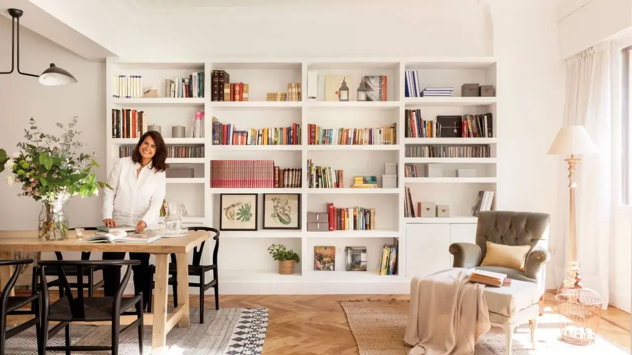 Una diseñadora casa sonríe junto a una mesa de comedor de madera, rodeada de estanterías llenas de libros y objetos decorativos.