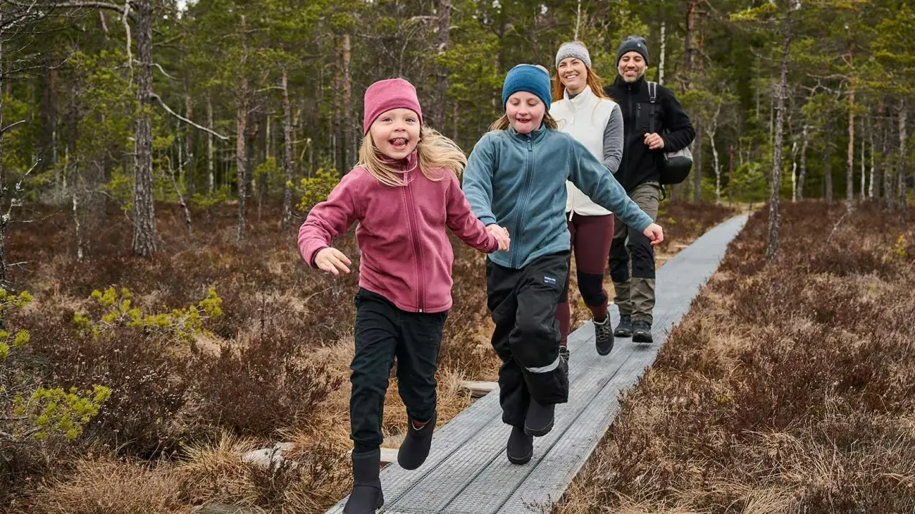 Familj njuter av aktiviteter i skogen med barn på en spång. Två barn springer glatt framför sina föräldrar i en vacker skogsmiljö.