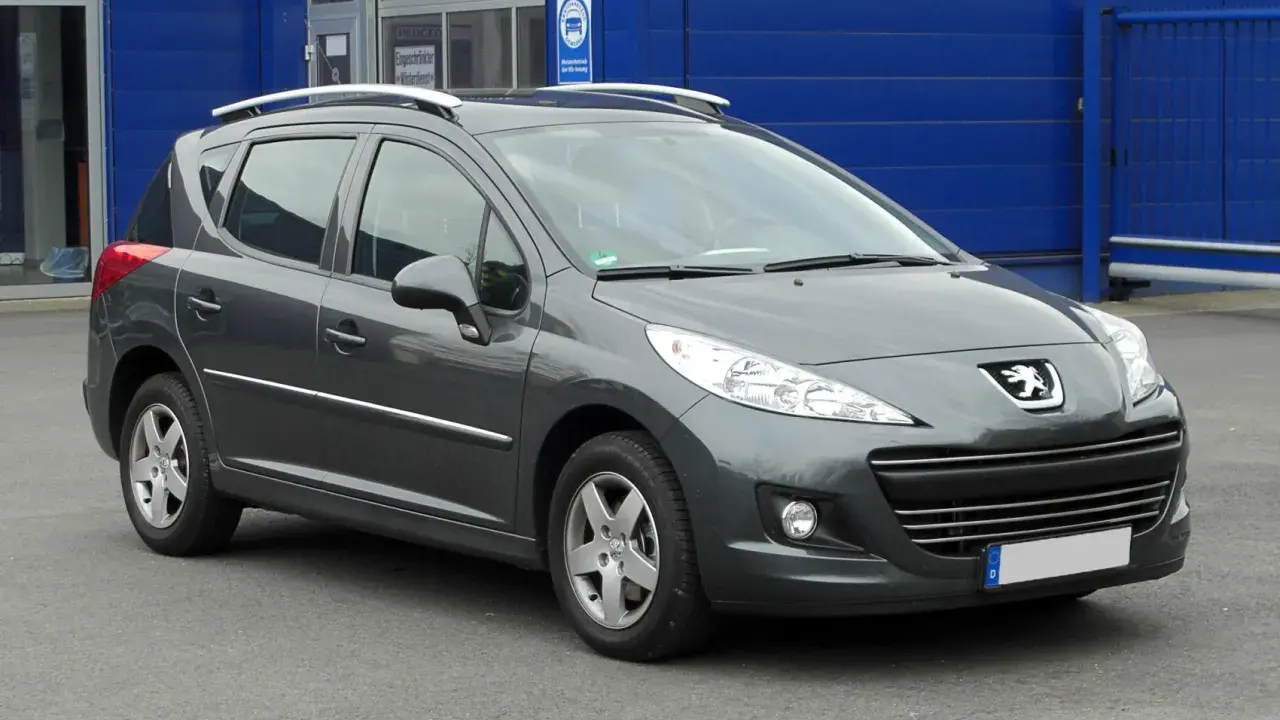 Szary Peugeot 207 1.4 benzyna z relingami dachowymi, zaparkowany przed niebieskim budynkiem.