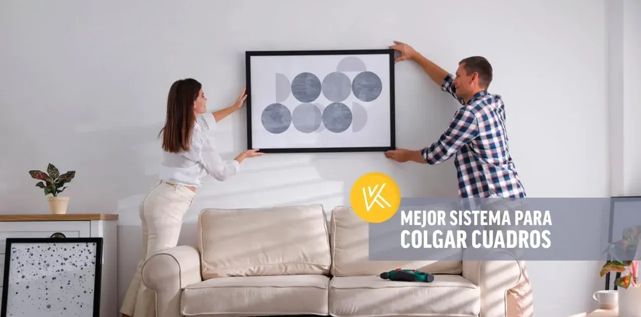 Pareja cuelga un cuadro moderno en pared de pladur. Un taladro descansa sobre el sofá, listo para la tarea.