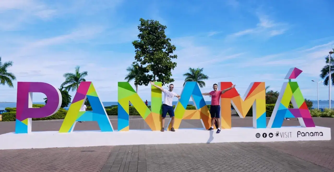 Two men pose with arms outstretched in front of a colorful "PANAMÁ" sign, suggesting Panama is gay friendly and a welcoming destination.