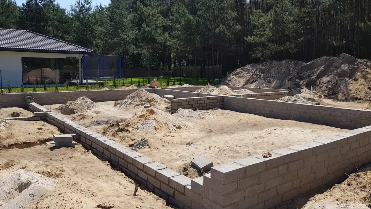 Fundamenty domu w budowie, piasek i stosy ziemi wokół. To etap samodzielnej budowy domu, widać postęp prac.
