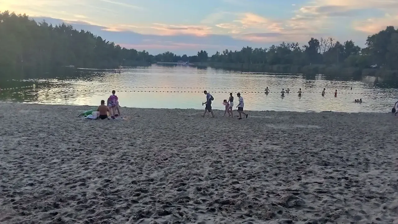Zachód słońca nad stawem w Jelczu Laskowicach. Ludzie spacerują po piaszczystej plaży i kąpią się w wodzie.