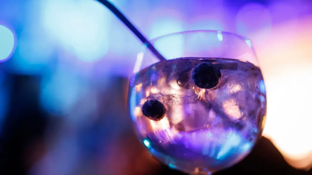 Ein Glas mit Eis und Beeren, in dem das tonic water leuchtet, vor einem verschwommenen, bunten Hintergrund.