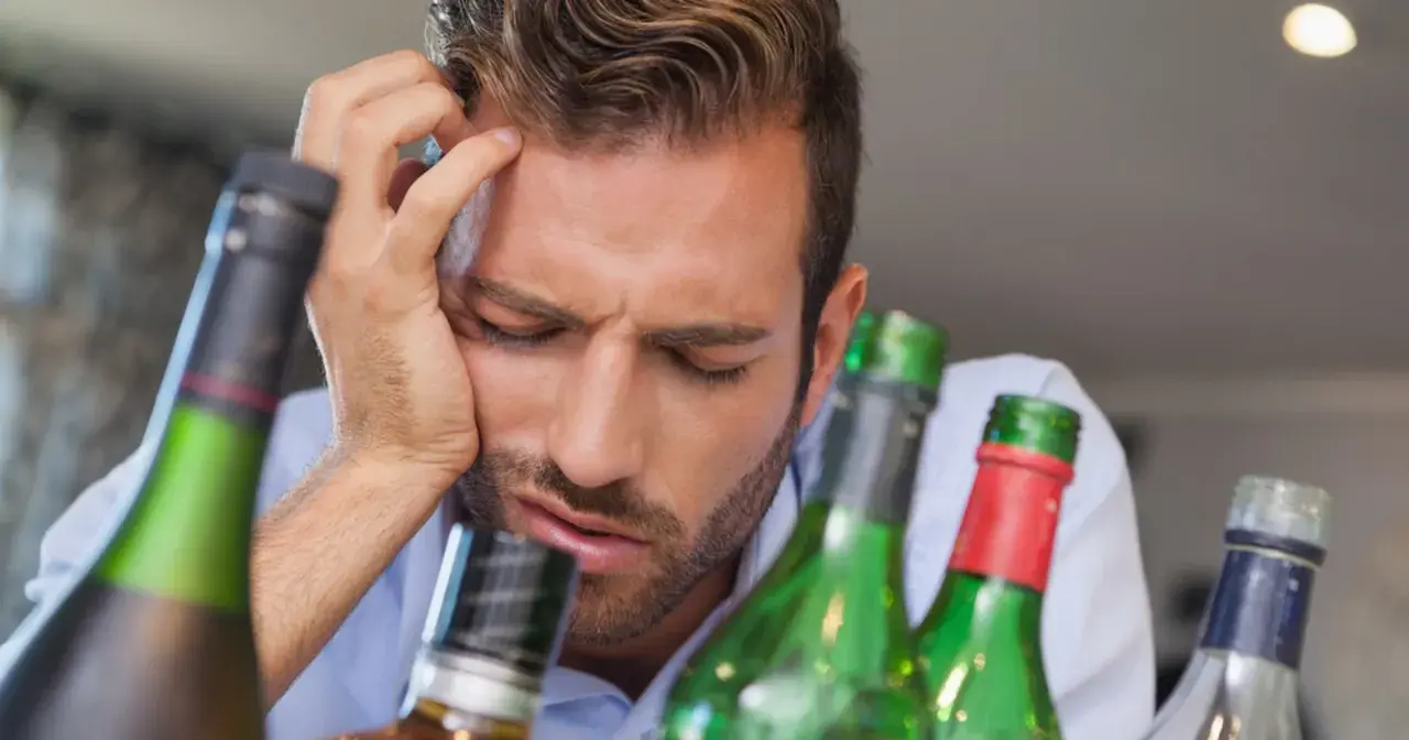 Mężczyzna z ręką na czole, otoczony butelkami, wyraźnie cierpi z powodu objawów przepicia alkoholowego.