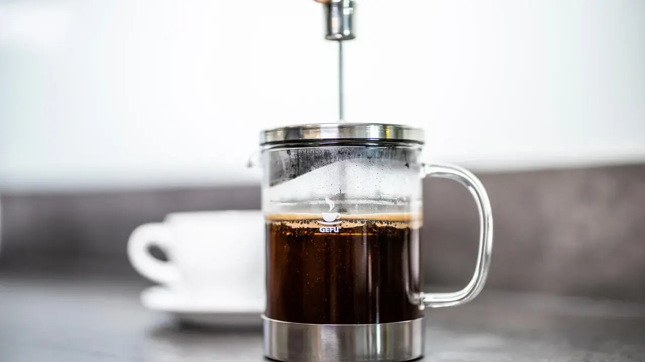 Eine Hand drückt den Stempel in eine French Press, um den perfekten Kaffee-Brew zuzubereiten. Eine Tasse wartet im Hintergrund.