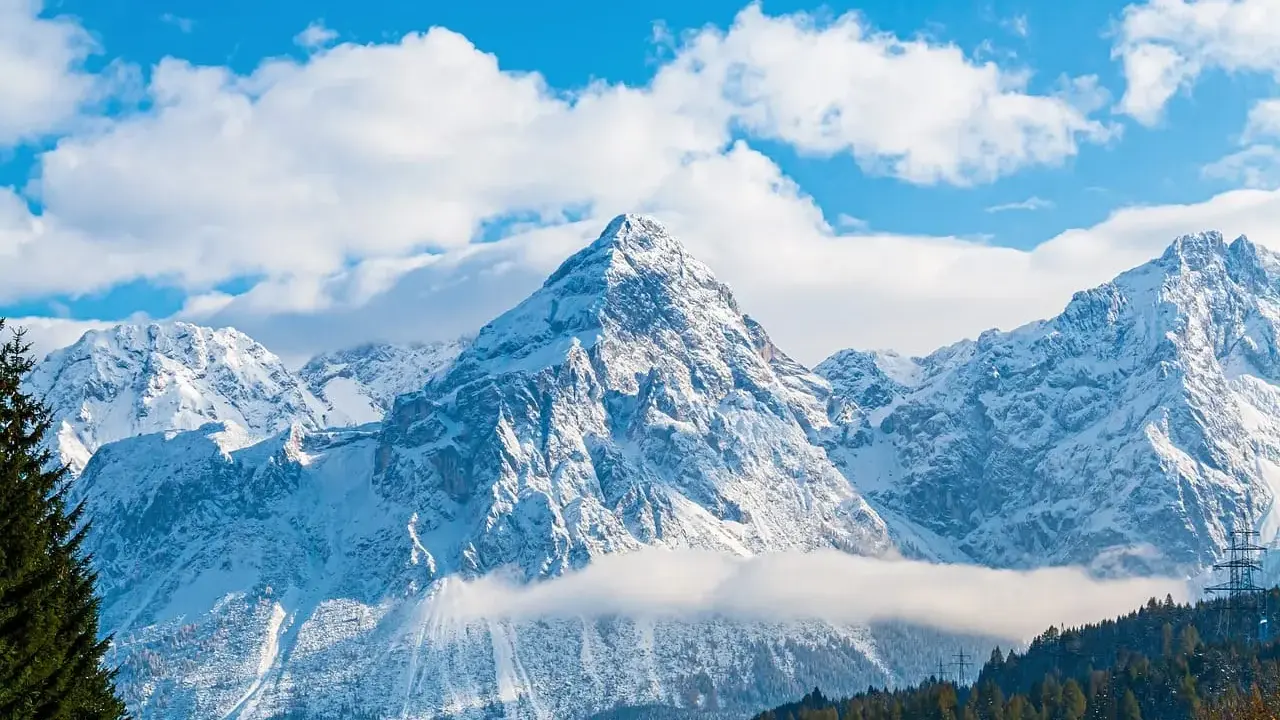 Majestatyczne, ośnieżone austria góry pod błękitnym niebem z chmurami. W dole las iglasty.