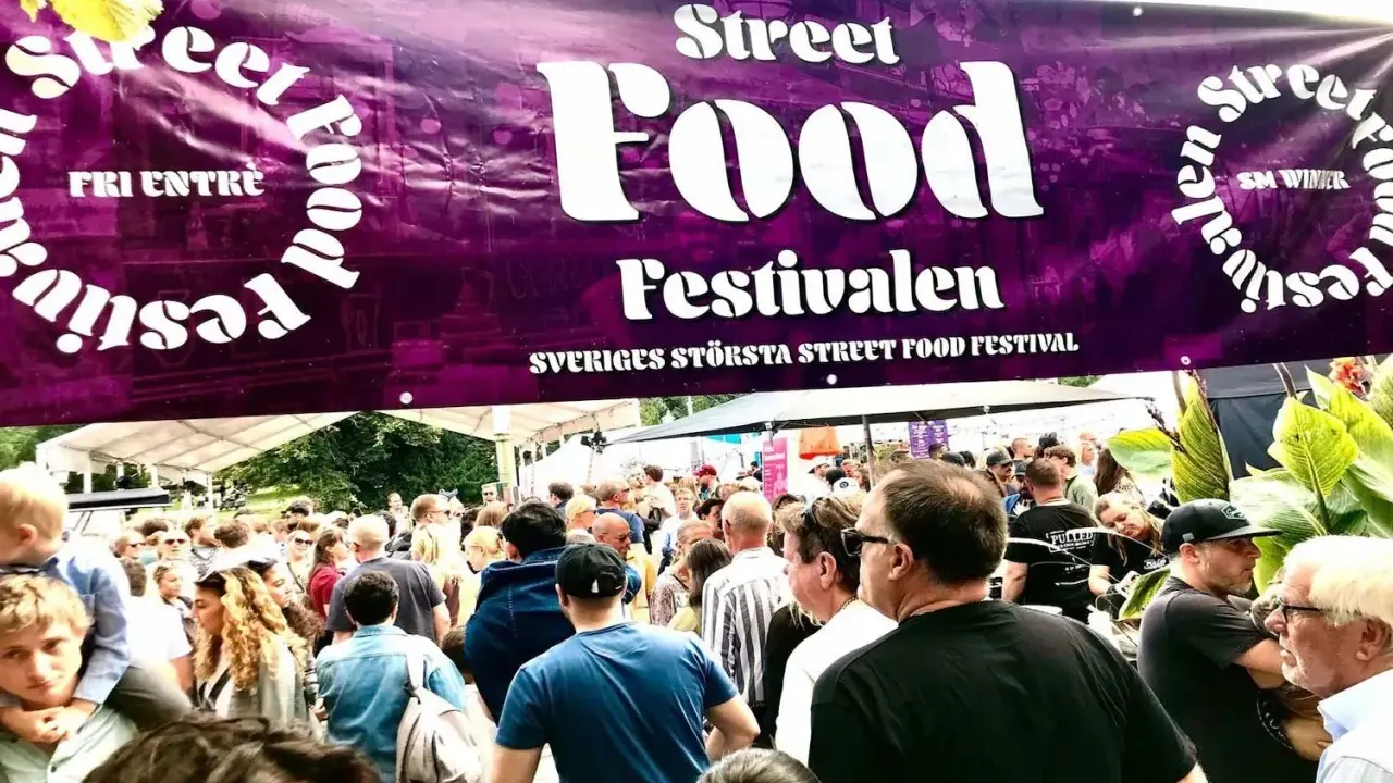Stor folkmassa vid Sveriges största street food festival. En lila banderoll med texten "Street Food Festivalen" hänger över besökarna.