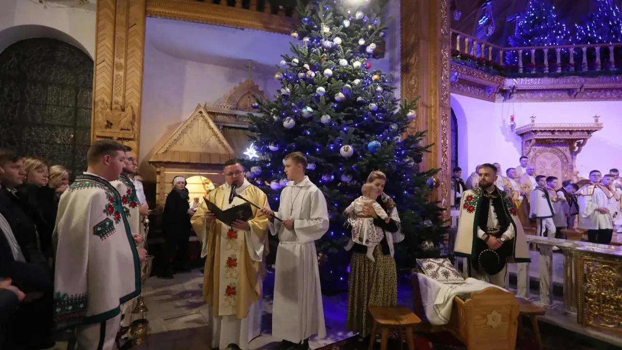 Pasterka w kościele w Zakopanem. Ksiądz czyta z księgi przy choince, obok ministranci i wierni w tradycyjnych strojach.