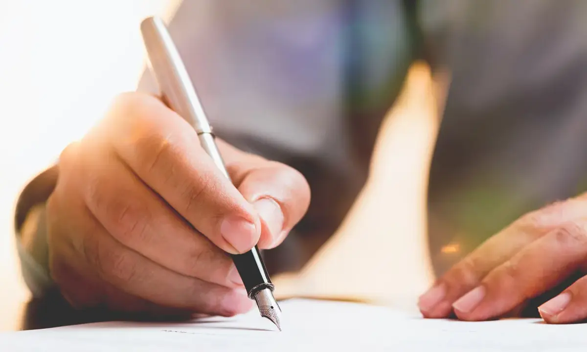 Dłoń pisze piórem po papierze, przygotowując dokument. Dowiedz się, jak napisać testament.