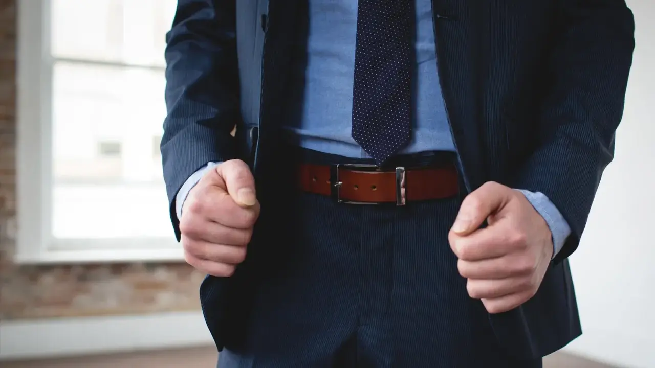 A man in a navy pinstripe suit and blue shirt wears a brown belt with black pants.
