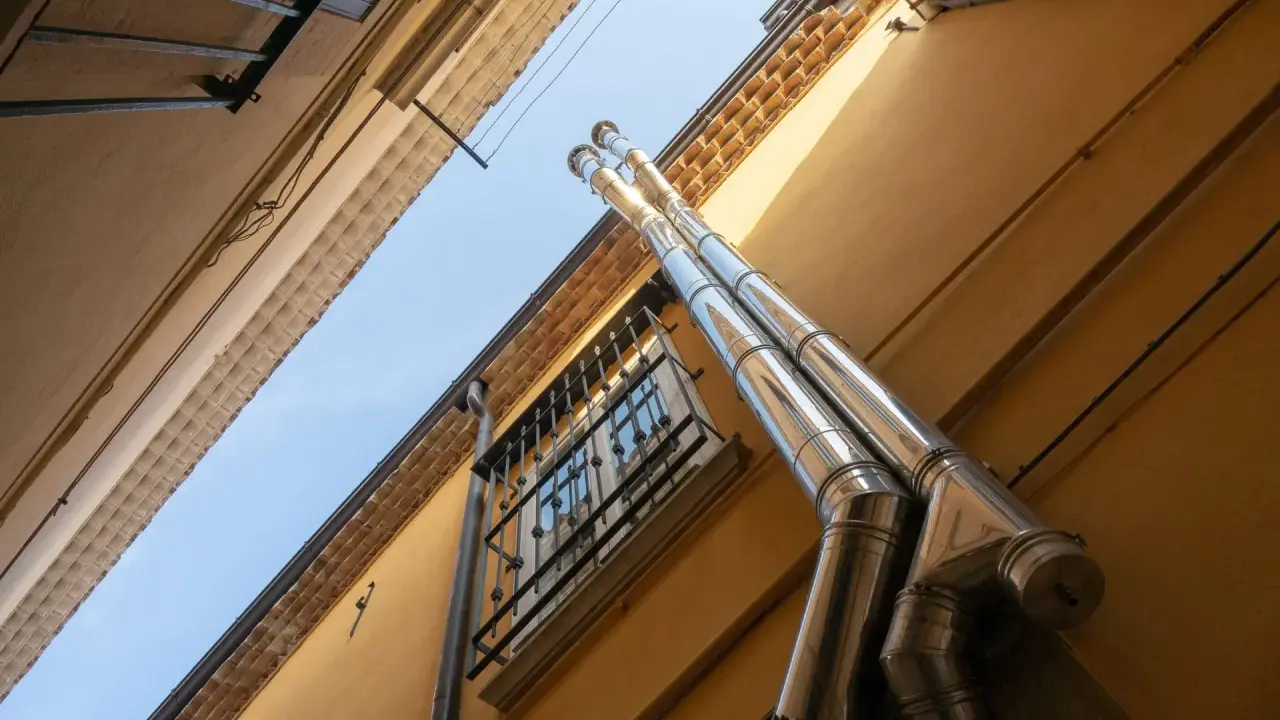 Dos tubos de salida humos estufa pellets brillan bajo el sol, ascendiendo por una pared amarilla junto a una ventana enrejada.