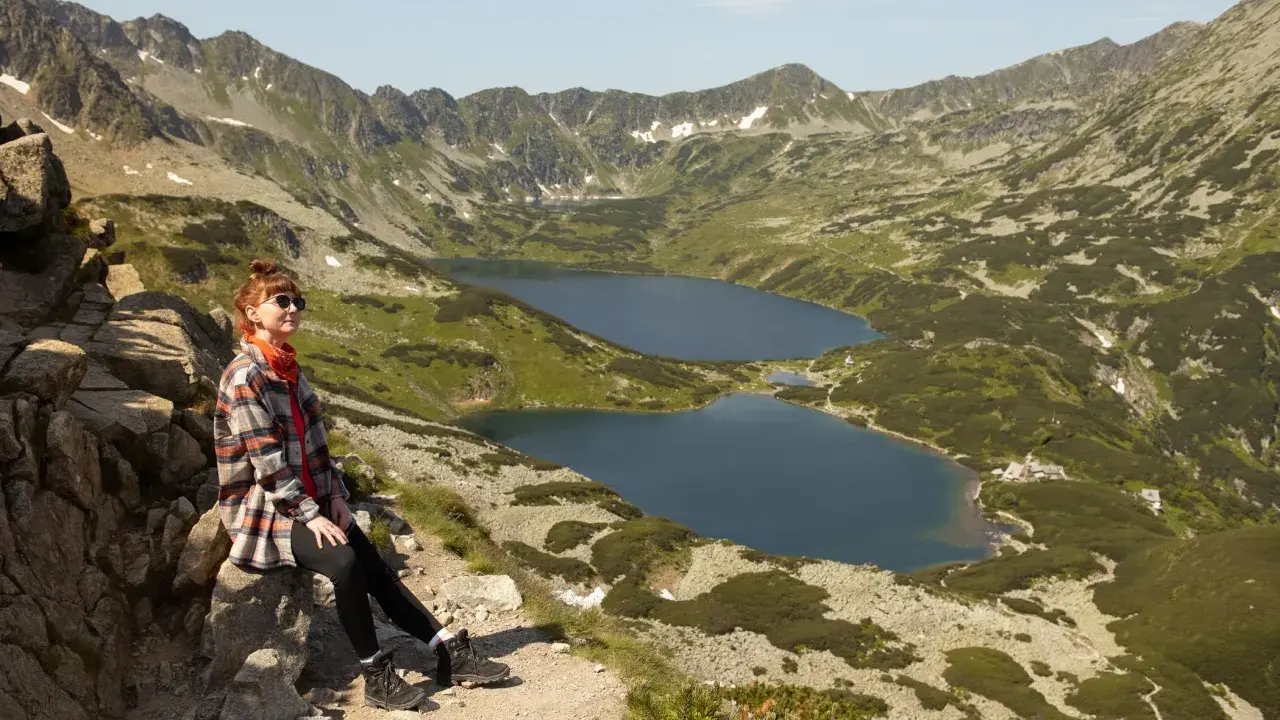 Kobieta odpoczywa na szlaku w Dolinie Pięciu Stawów. W tle rozciągają się góry i jeziora.