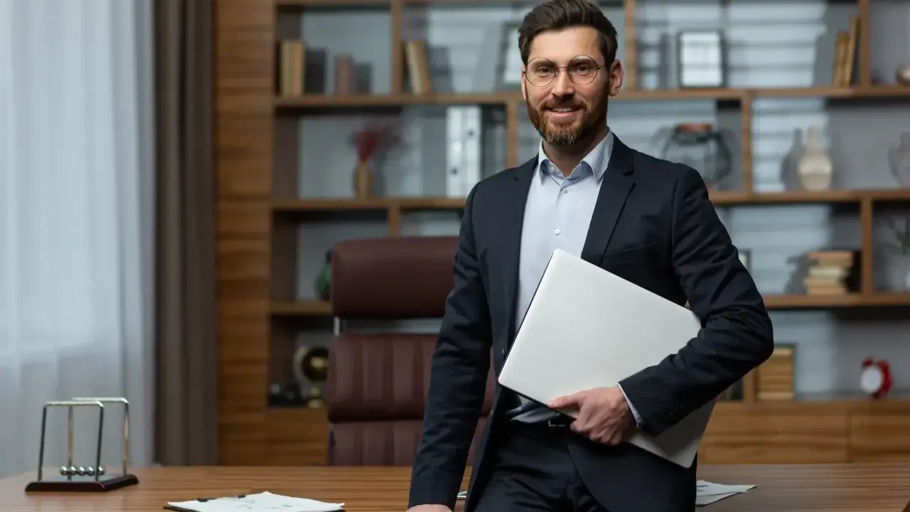 Uśmiechnięty dyrektor szkoły w garniturze, trzymający laptopa, stoi przy biurku w gabinecie z regałami pełnymi książek.