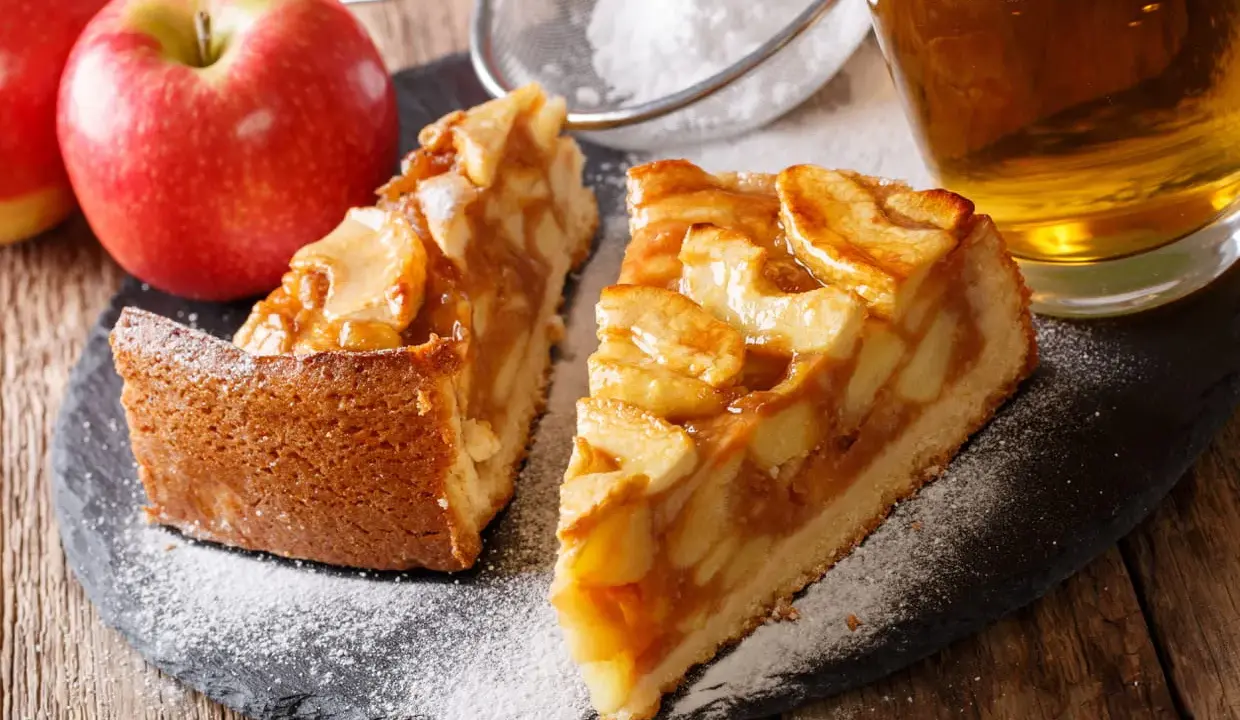 Deux parts de tarte aux pommes sur une ardoise, saupoudrées de sucre glace, avec des pommes et un verre de cidre.