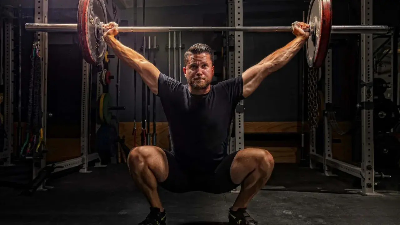 Ein Mann im tiefen Squat mit einer Langhantel über dem Kopf. Seine Muskeln sind angespannt, bereit für den nächsten Schritt im **power lifting**.