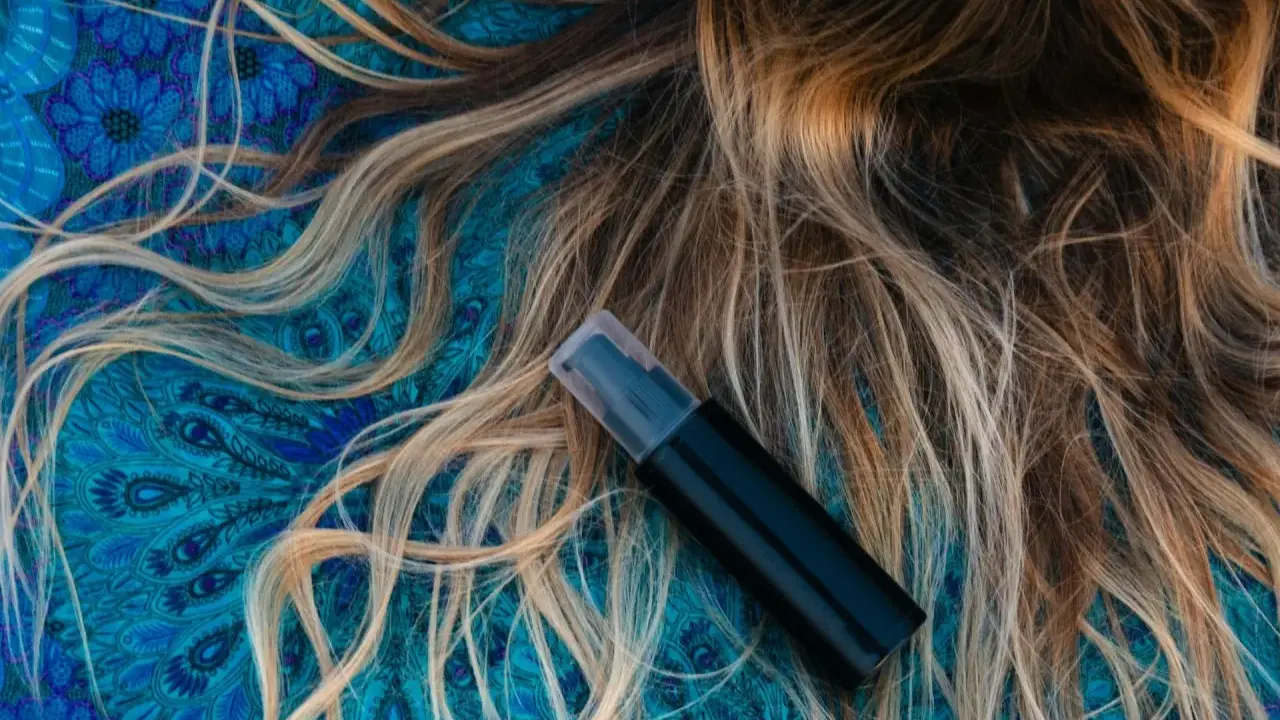 Czarny atomizer na tle błękitnego wzoru, obok rozpuszczonych, jasnych włosów. Idealny sposób, jak nawilżyć włosy.