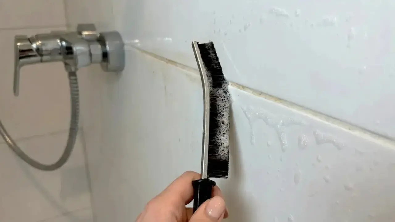 Hand reinigt Duschfugen mit einer Bürste und Schaum.