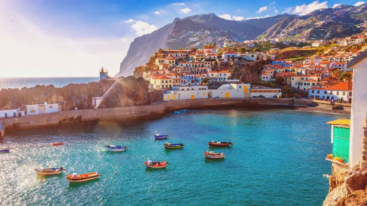 Bunte Häuser schmiegen sich an steile Hänge auf den Inseln bei Madeira. Kleine Boote schaukeln im azurblauen Wasser vor einem Leuchtturm.