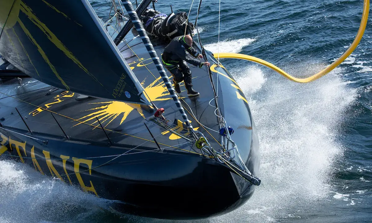 Un skipper manœuvre sur l'IMOCA Bureau Vallée, le bateau noir et jaune fend les vagues dans un embrun spectaculaire.