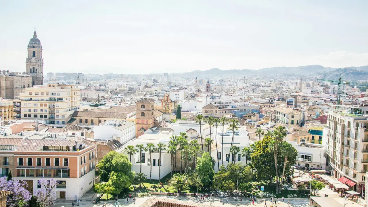Blick über die Dächer von Malaga, mit dem markanten Glockenturm der Kathedrale und Palmen. Der malaga hafen ist in der Ferne zu erahnen.