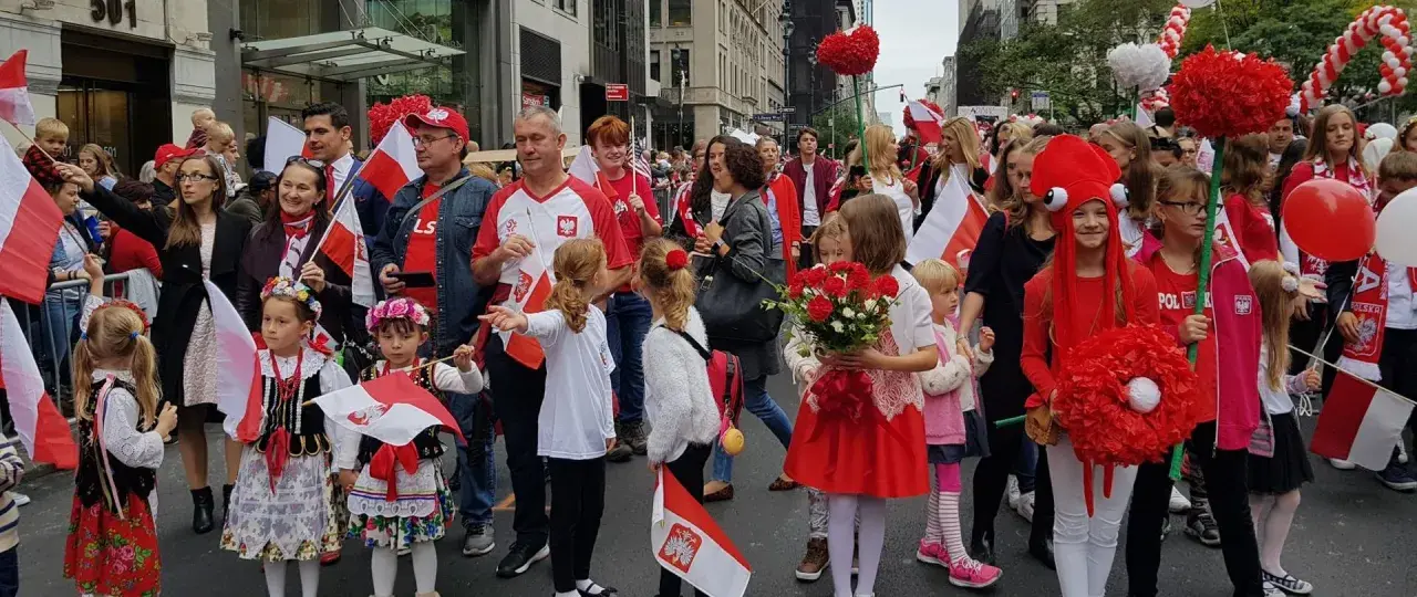 Dzieci i dorośli w strojach ludowych i z polskimi flagami maszerują ulicą. Widać serca z czerwonych balonów i kwiatów. Polacy w Szwecji świętują.
