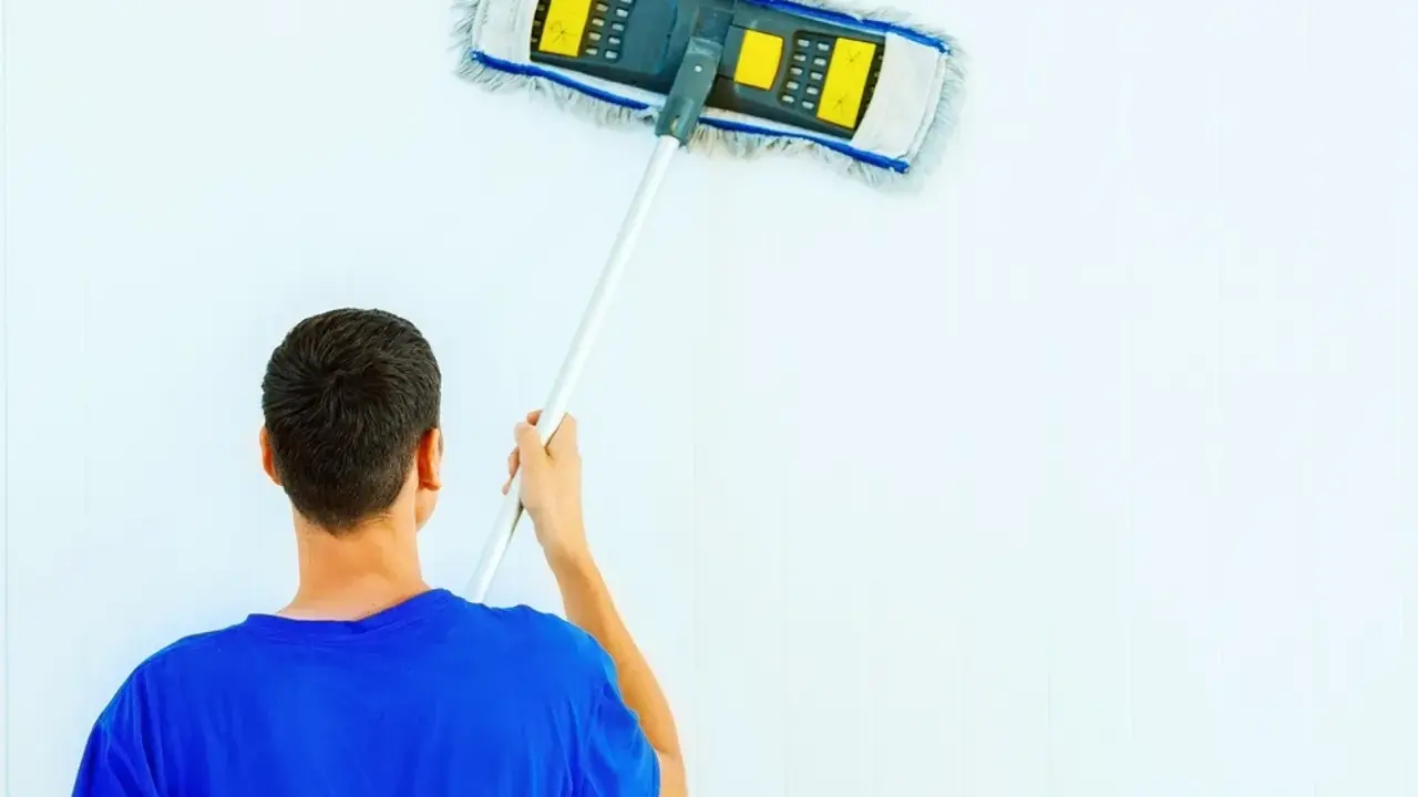 Hombre con camiseta azul limpiando paredes blancas con un plumero.