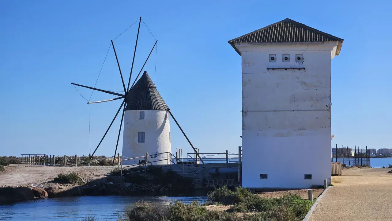 Altes Windrad und ein weißes Gebäude am Mar Menor, mit Wasser und Vegetation im Vordergrund.