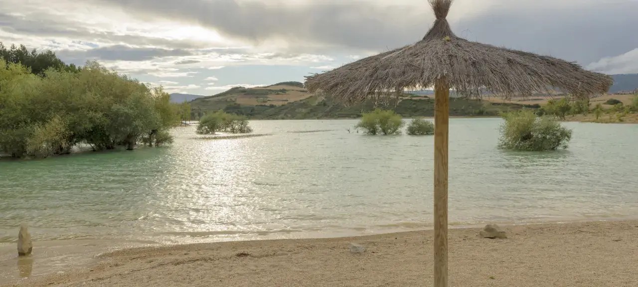 Sombrilla de paja en la orilla del pantano de Alloz. El agua turquesa, árboles emergiendo y colinas al fondo invitan a descubrir cómo llegar.