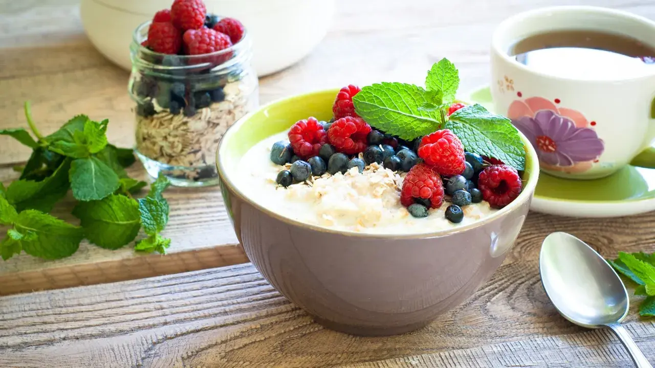 Schüssel mit Haferflocken mit Kefir, garniert mit Himbeeren, Blaubeeren und Minze. Daneben ein Glas Müsli und eine Tasse Tee. Gesundes Frühstück.