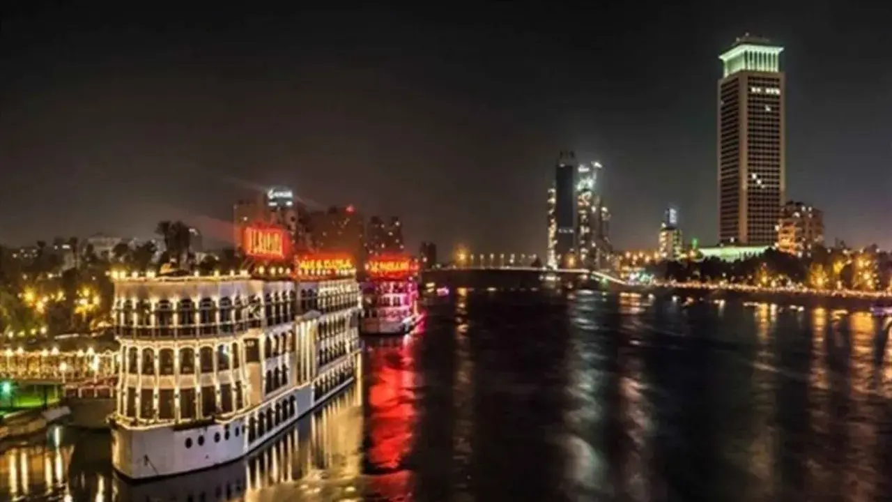 Beleuchtete Nilschiffe und moderne Hochhäuser spiegeln sich im Wasser. Ein magischer Anblick von Kairo bei Nacht.