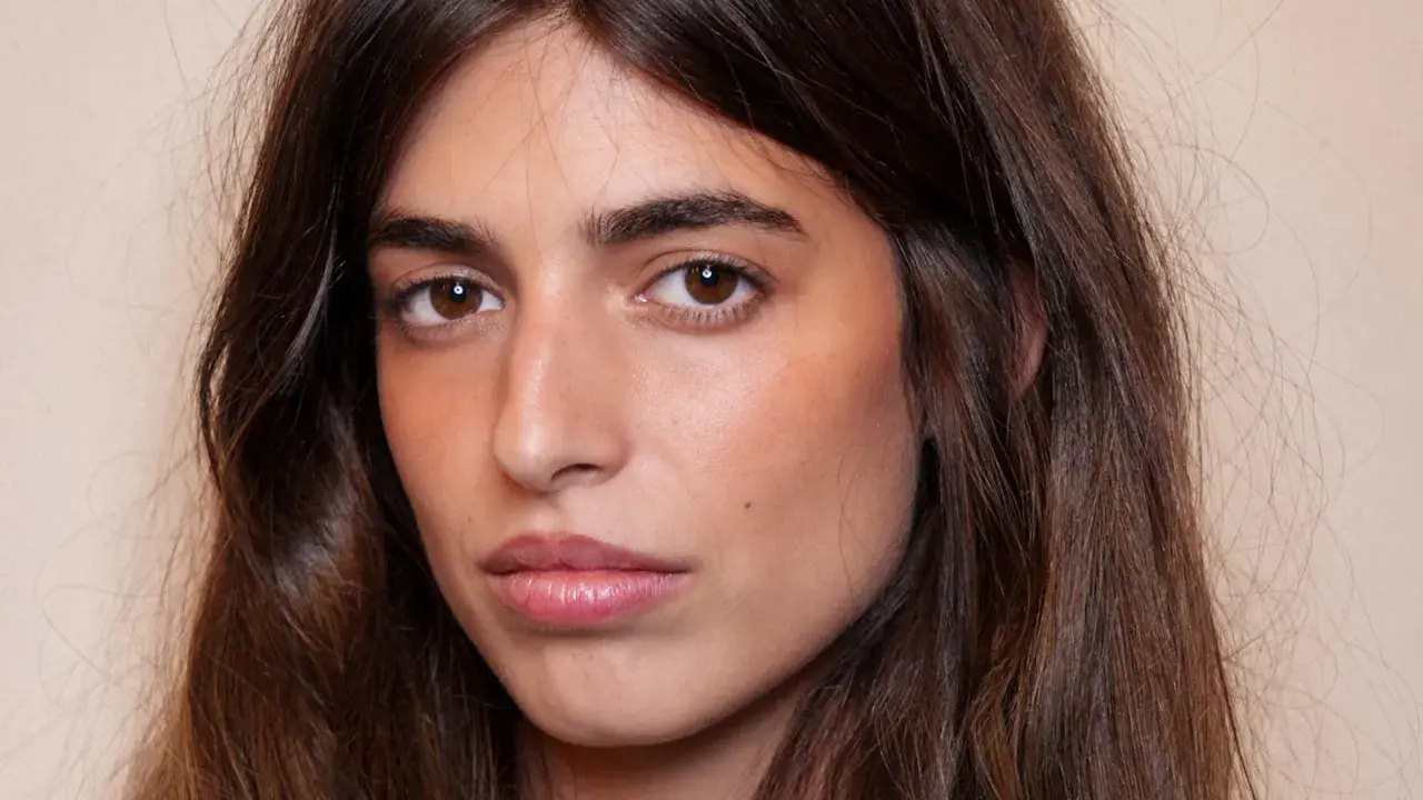 Portrait d'une jeune femme aux longs cheveux bruns. Son maquillage met en valeur ses yeux et ses lèvres, avec un soin particulier apporté à son sourcil poil à poil.