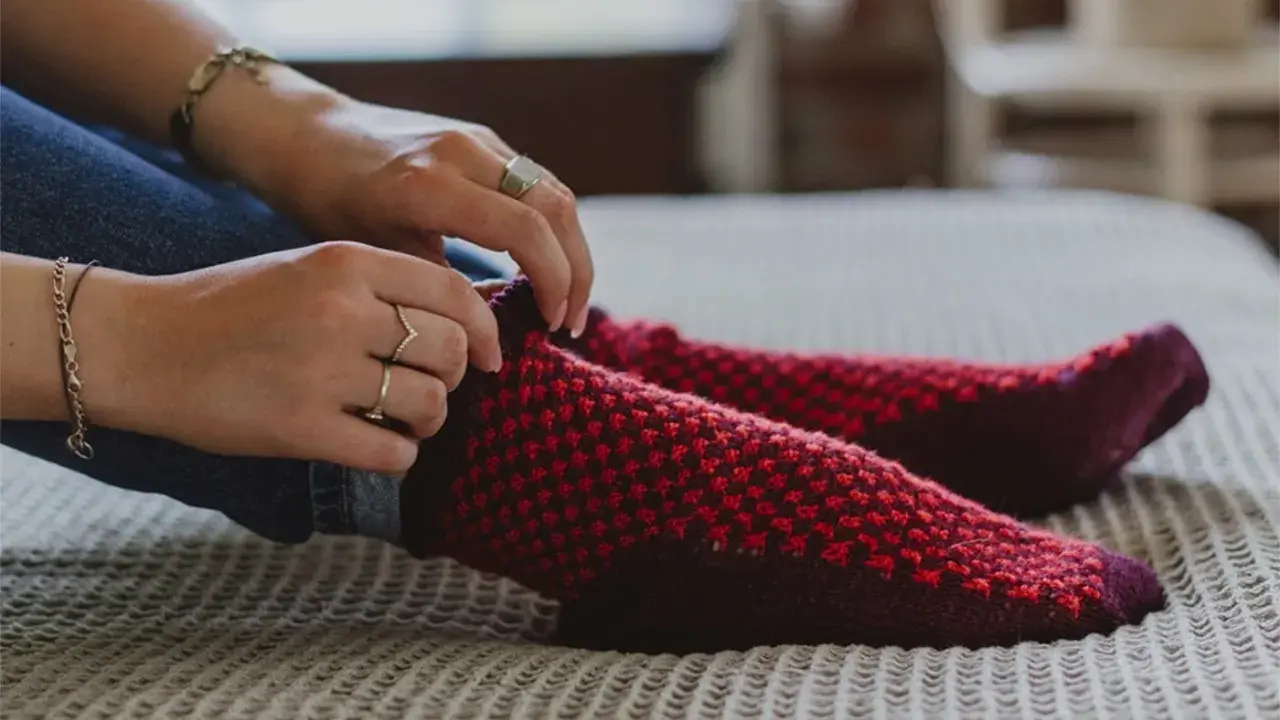 Manos ajustando calcetines rojos y morados tejidos a mano con agujas circulares.