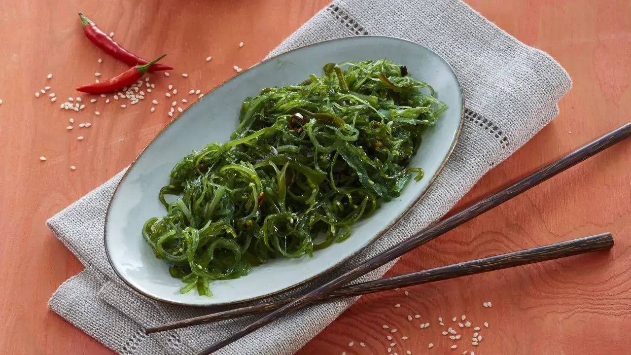 Ein Teller mit leckerem Wakame Salat, garniert mit Chilis und Sesam. Dazu liegen Holzstäbchen bereit.