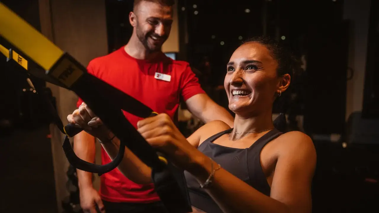 Frau lacht und trainiert mit TRX-Bändern, ihr Trainer lächelt. Sie will schnell wieder fit werden.