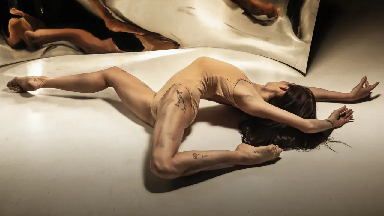 Młoda kobieta w cielistym body wygina się w łuk na podłodze, jej odbicie w lustrze tworzy surrealistyczny obraz. Jak z opowiadanie erotyczne nastolatki.