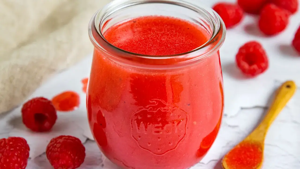 Ein Glas mit leuchtend roter Himbeer-Vinaigrette, umgeben von frischen Himbeeren und einem Holzlöffel mit etwas Vinaigrette.
