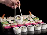 Dłoń z pałeczkami podnosi sushi. Różnorodne rolki, w tym zielone i różowe, czekają na spróbowanie. Nauka jak jeść pałeczkami to klucz do tej uczty.