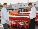 Dwóch mężczyzn ogląda czerwoną maszynę rolniczą, prawdopodobnie siewnik lub bronę, na tle innych maszyn i budynków.